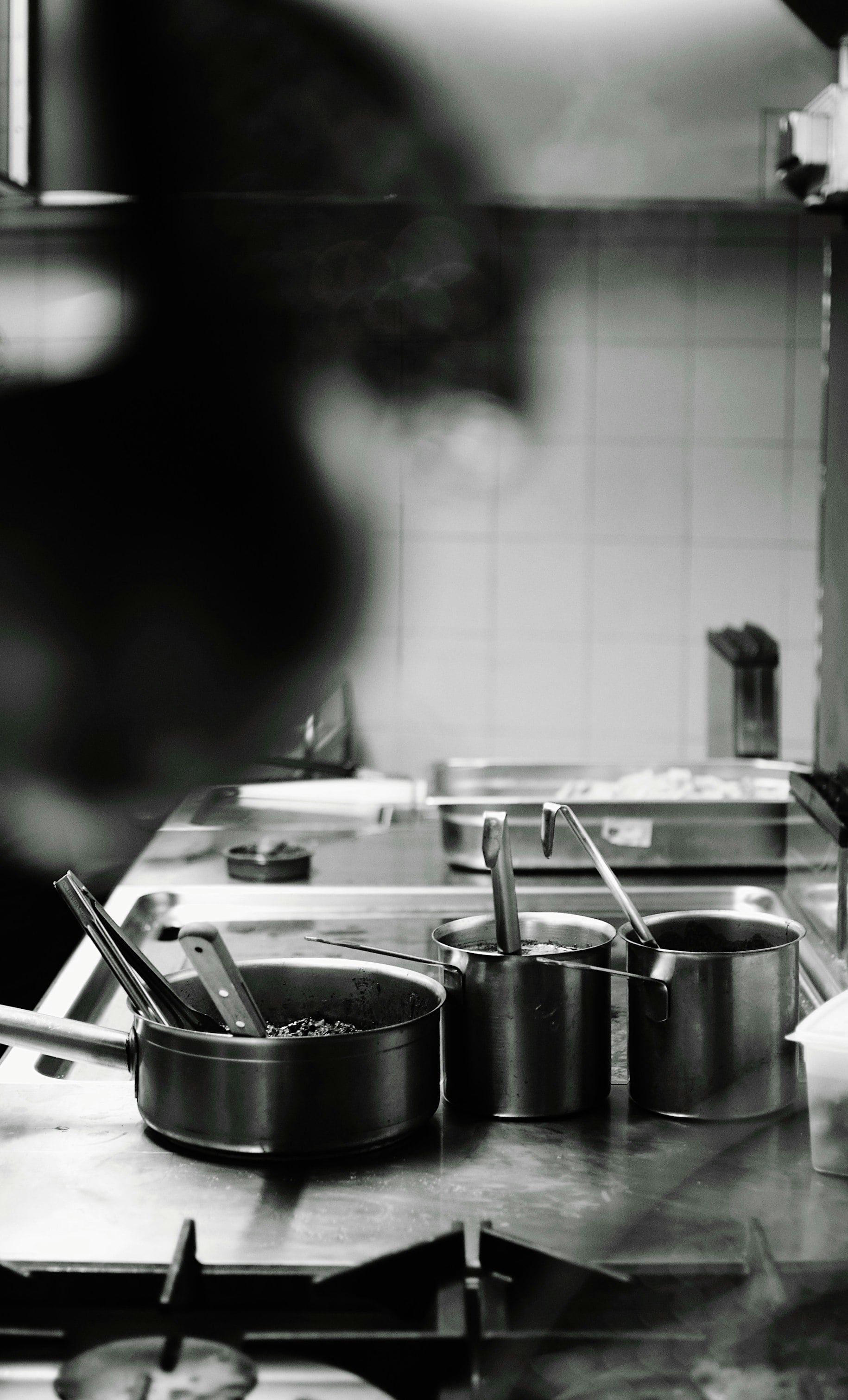 Vista en blanco y negro de una cocina de restaurante con cazos de acero, cucharones y pinzas, y un cocinero desenfocado en primer plano a la izquierda