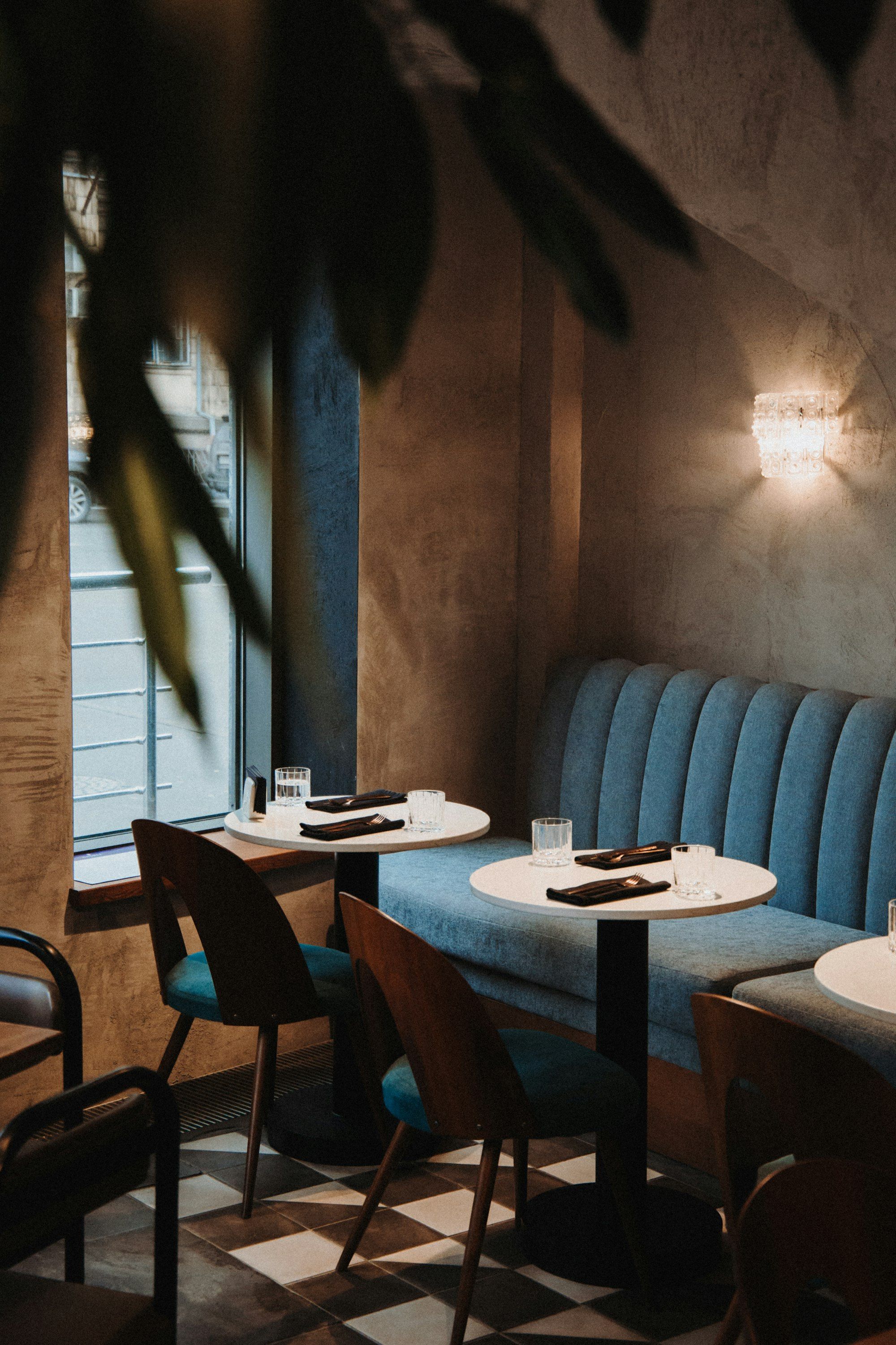 Comedor con bancos de terciopelo azul, mesas redondas blancas puestas para cenar y suelo de cuadros, con luz cálida