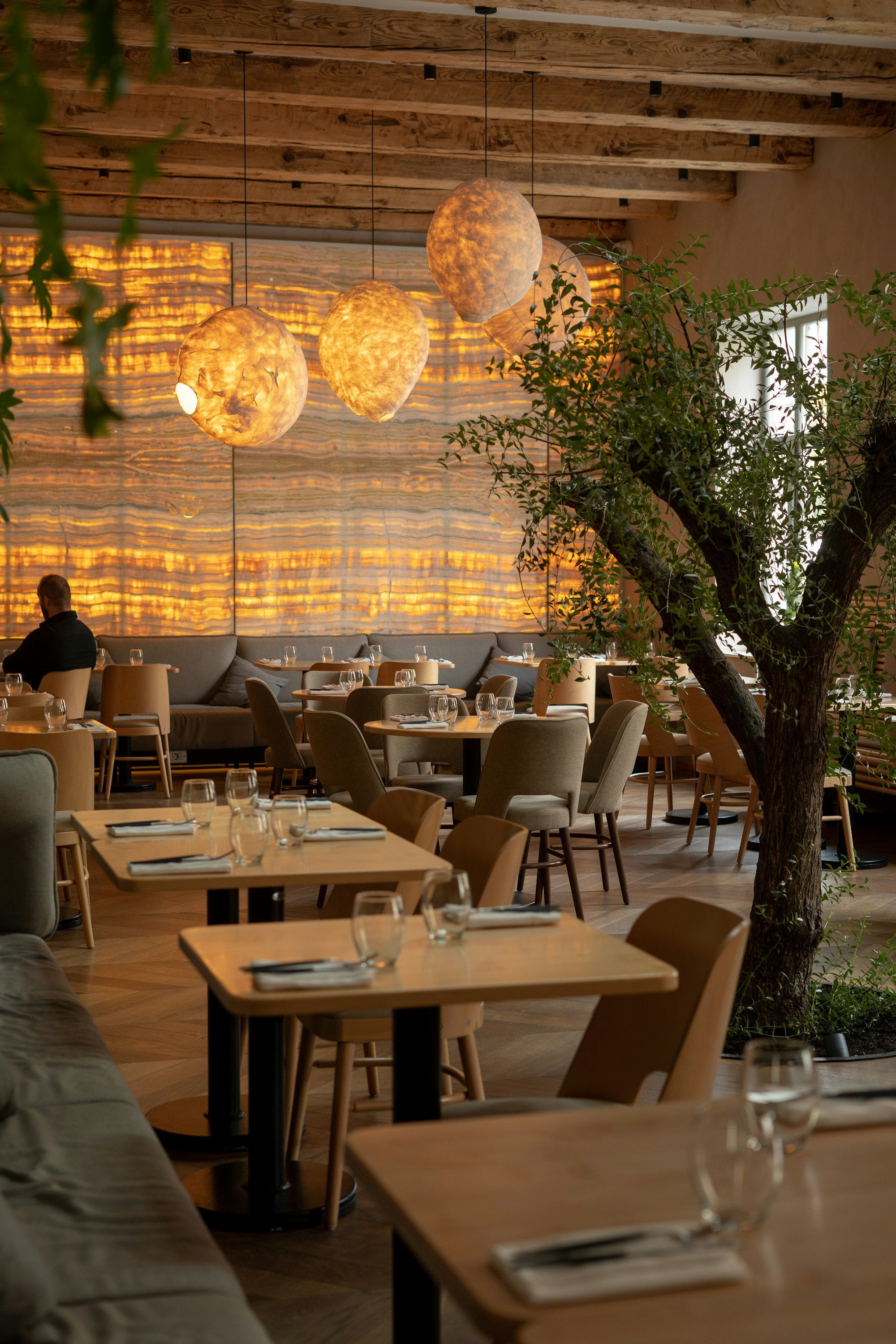 Comedor cálido con una pared de ónice iluminada en ámbar, lámparas colgantes, mesas y un árbol en medio de la sala