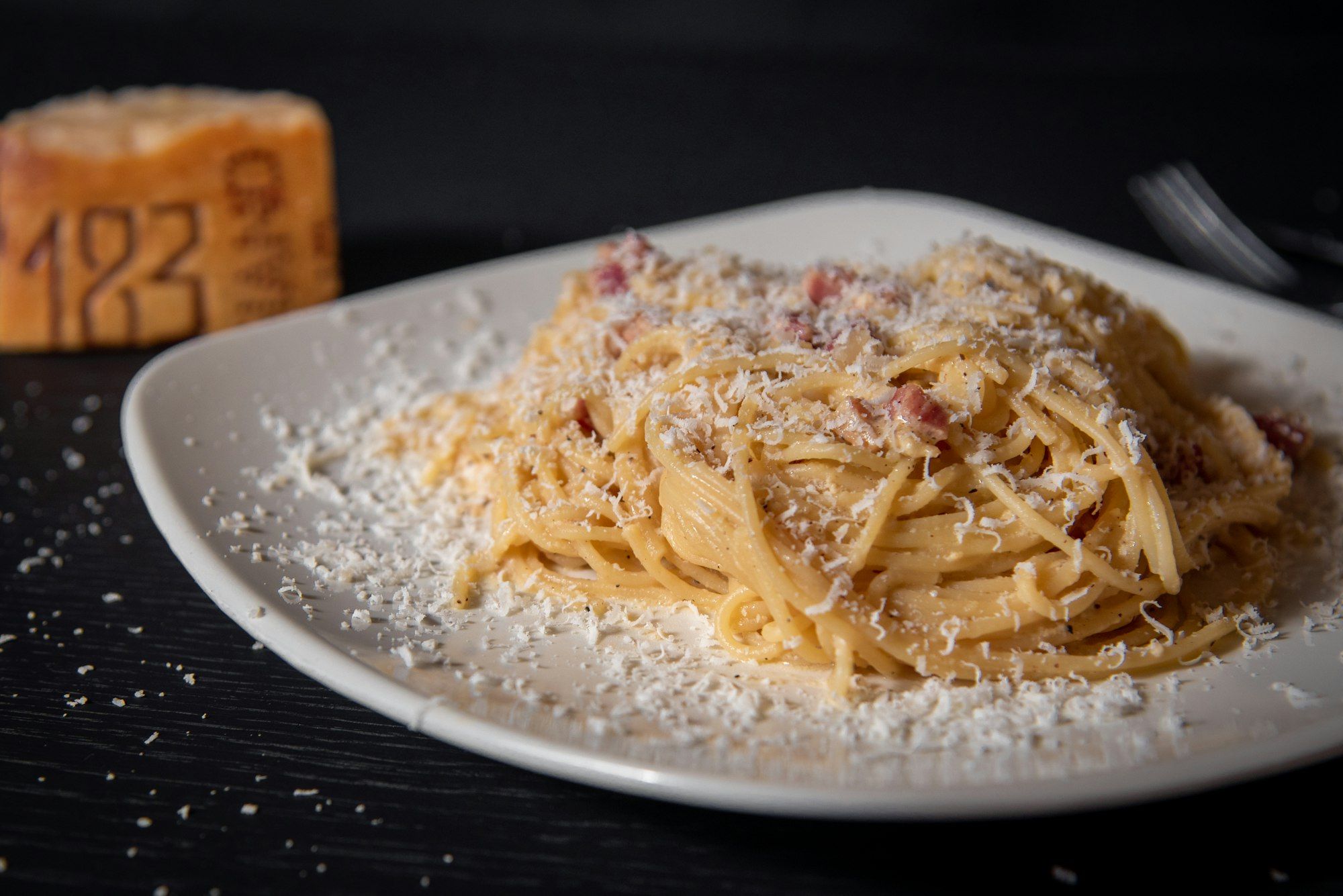 Espaguetis a la carbonara en un plato blanco sobre fondo oscuro, con una rueda de parmesano al fondo